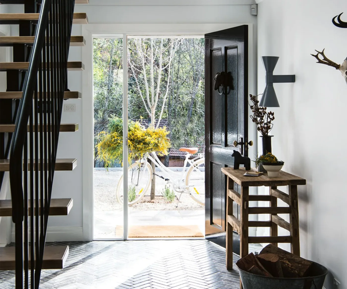 Country entrance with an open dark front door, glazed side panel, wooden side table, dark stair railing and herringbone patterned floor.