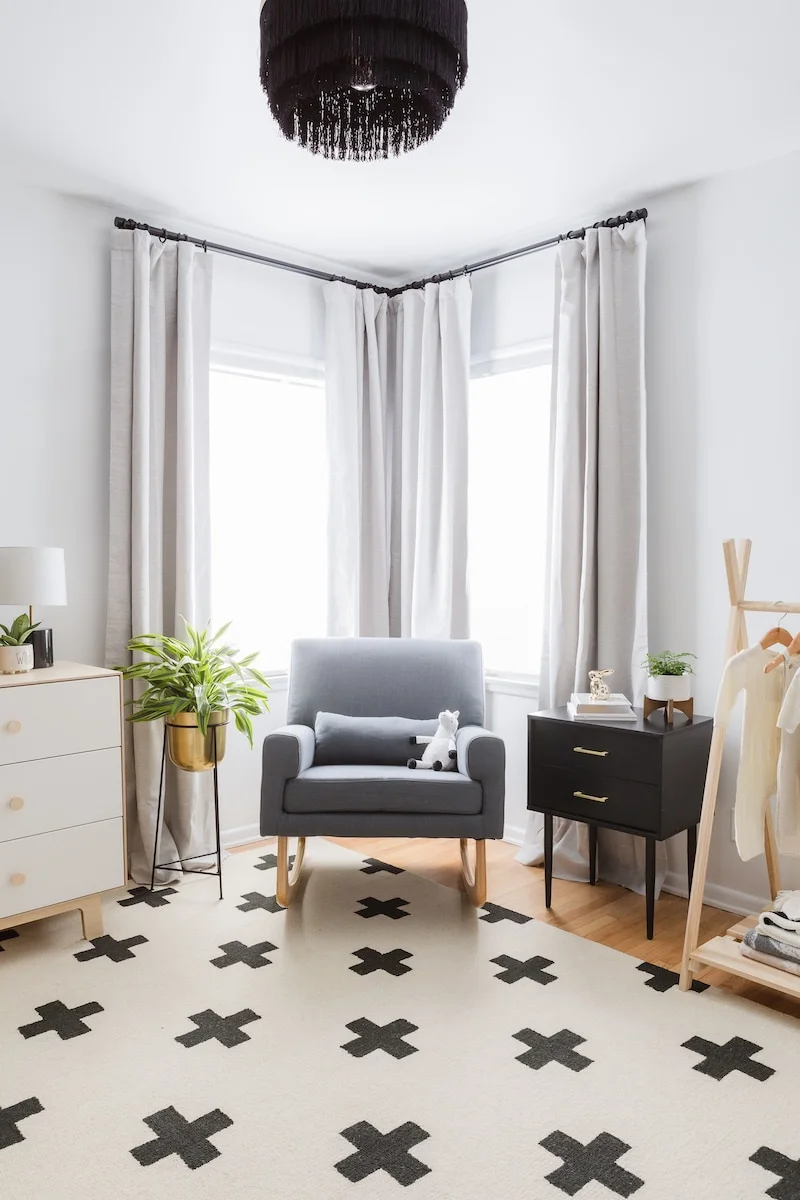 A gray nursery adult chair sits beside dark drawer storage and a pale dresser between two curtained windows above a pale rug with dark crosses
