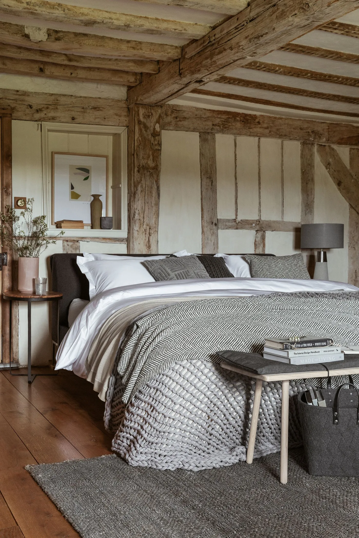 Country bedroom with exposed wooden wall and ceiling timbers, a dark headboard, pale bedding, gray textured throw and a small round bedside table.
