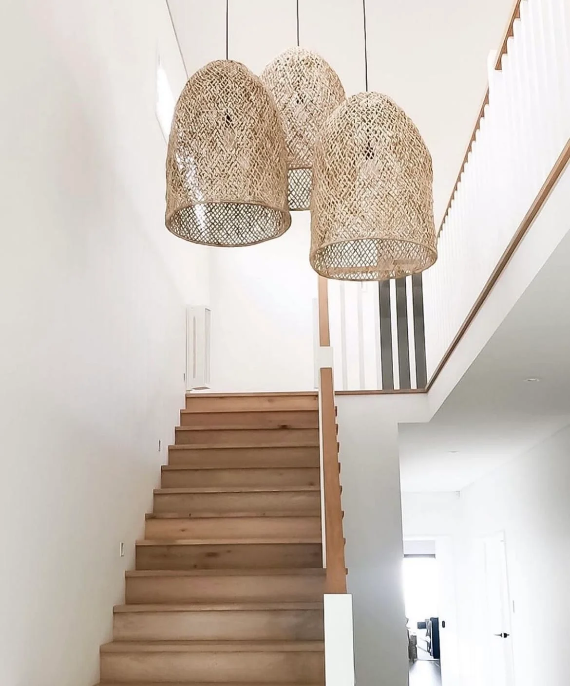 Three woven pendant shades hang over a wooden staircase with white walls and wooden handrails