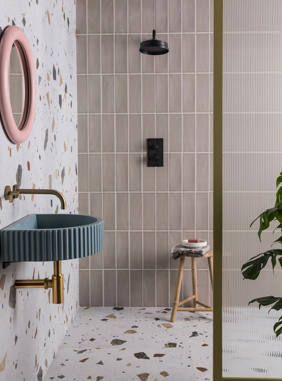 A ribbed blue basin sits beneath a pink round mirror beside speckled walls and floor, pale pink shower tiles and black fittings