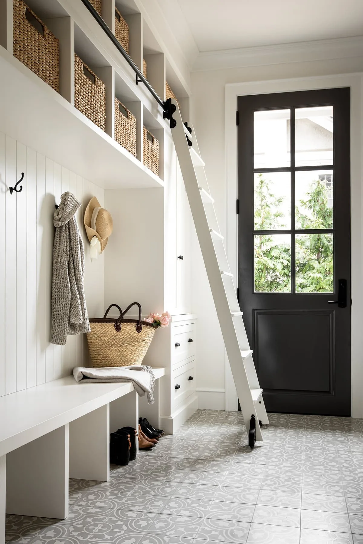 White mudroom bench and paneled cubbies with high woven baskets, a ladder and a black glazed door.