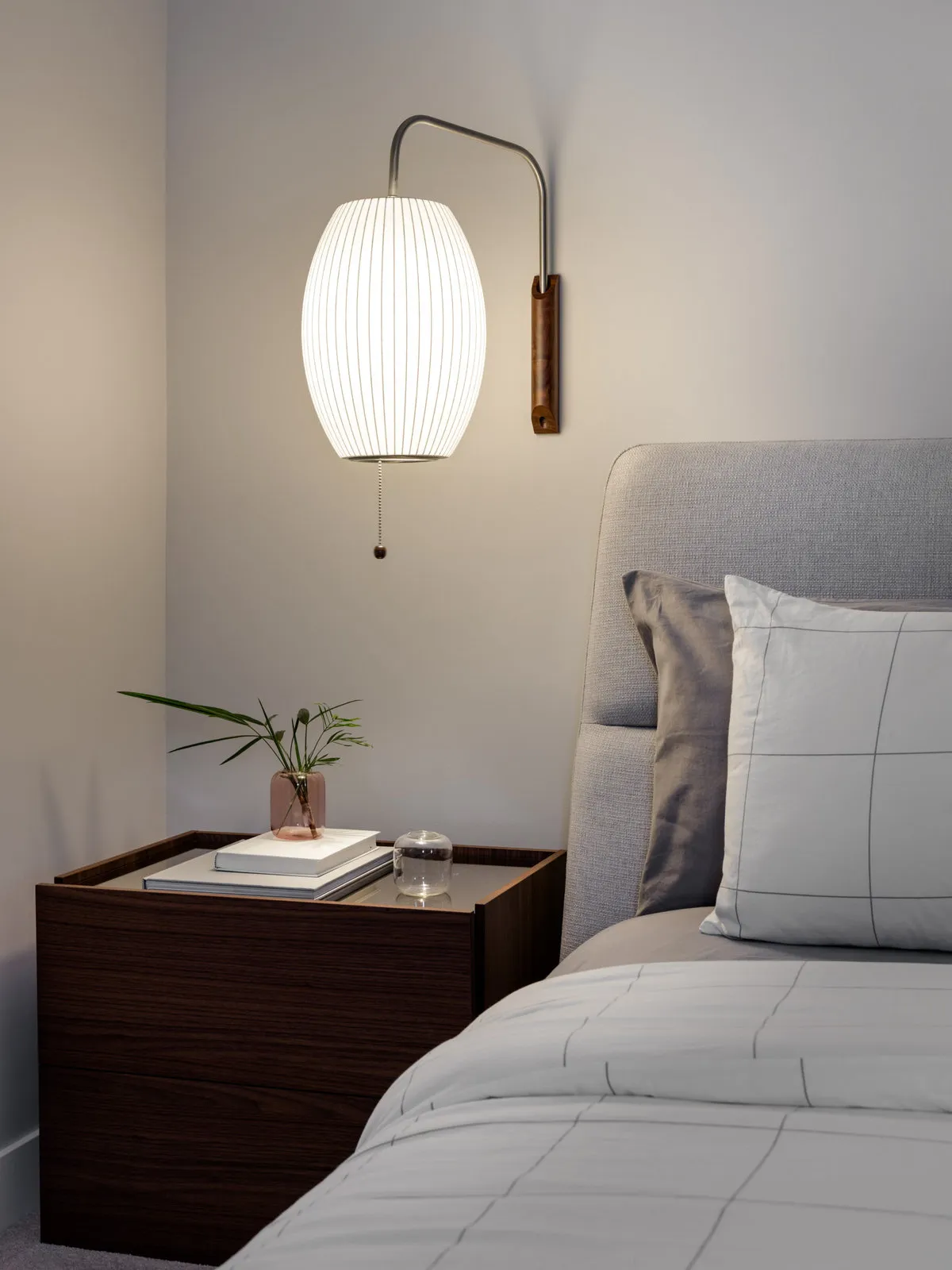 A pale pleated wall light hangs from a curved arm above a timber bedside table beside an upholstered headboard.