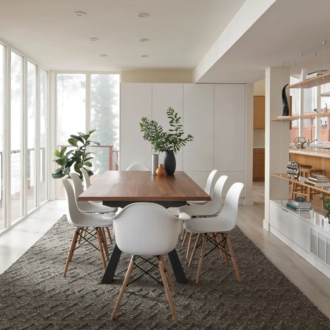 Pale open display shelves beside a wood dining table and white chairs in a bright windowed room