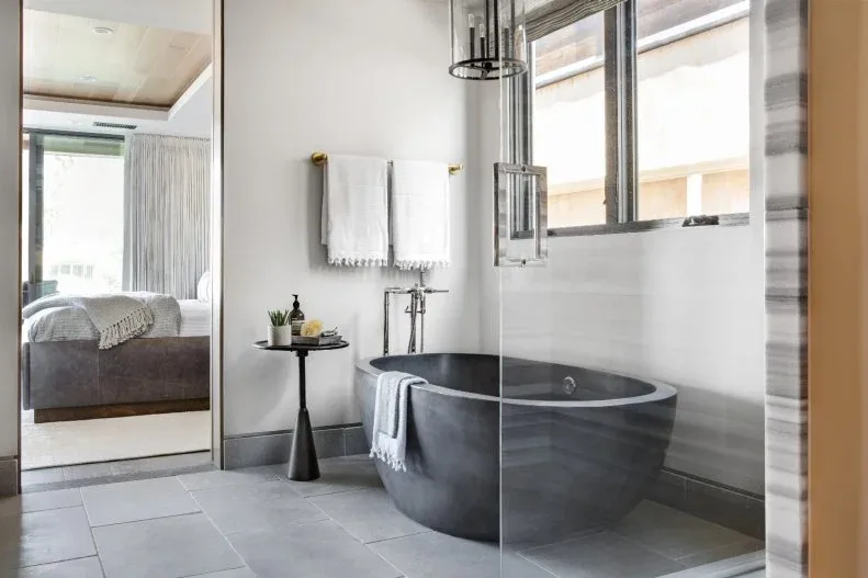 Dark gray rounded freestanding bath beside a clear glass screen, gray floor, white wall, broad window, hanging fixtures, and black pedestal table.