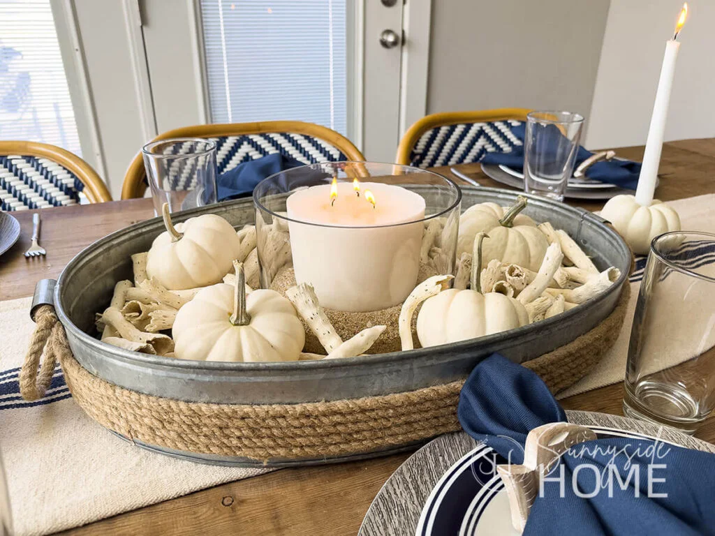 White pumpkins, pale branching ornaments and a candle bowl sit in an oval rope-edged tray above a blue-and-white striped runner