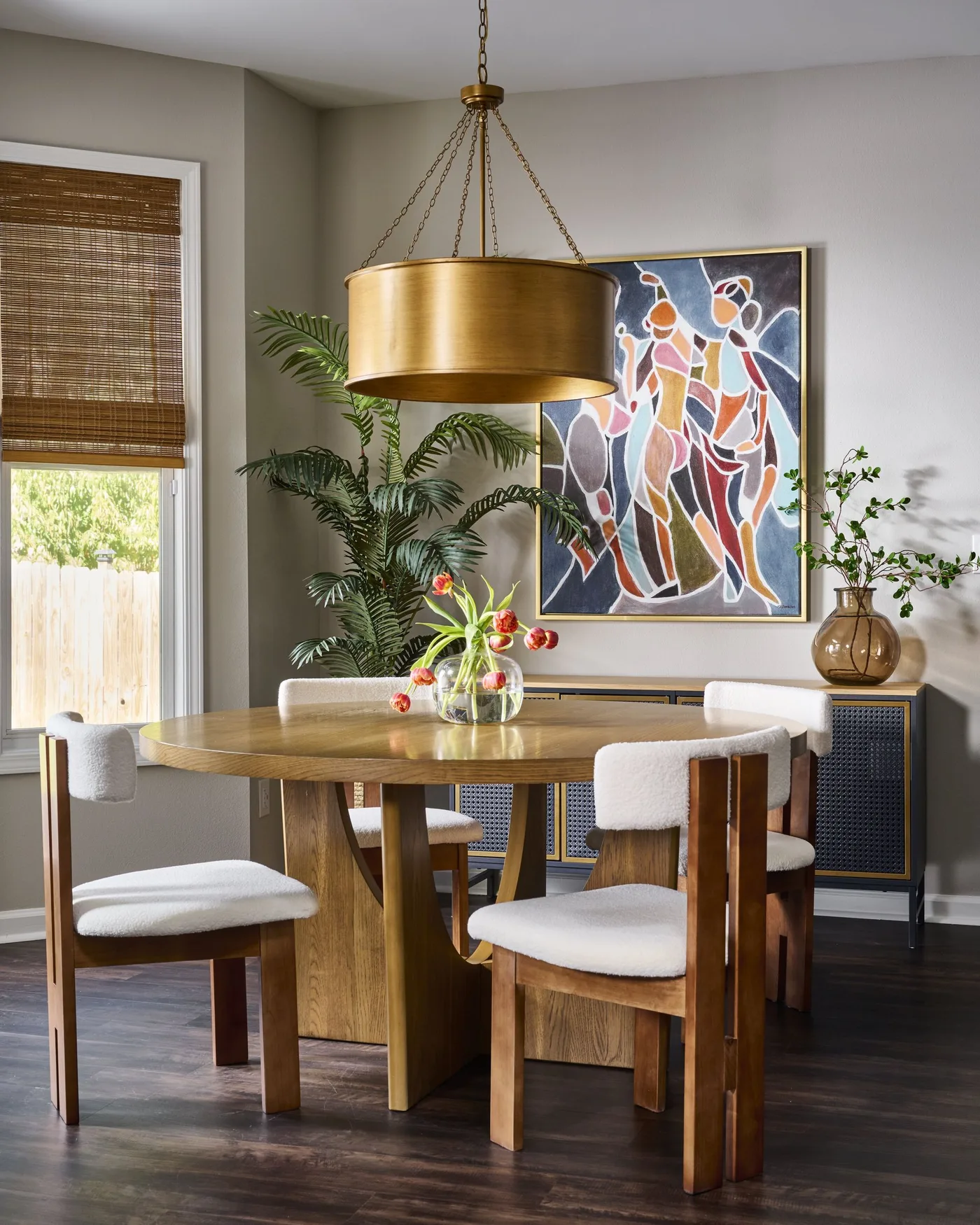 Gold-toned drum light above a round wood table with pale upholstered chairs and colorful artwork