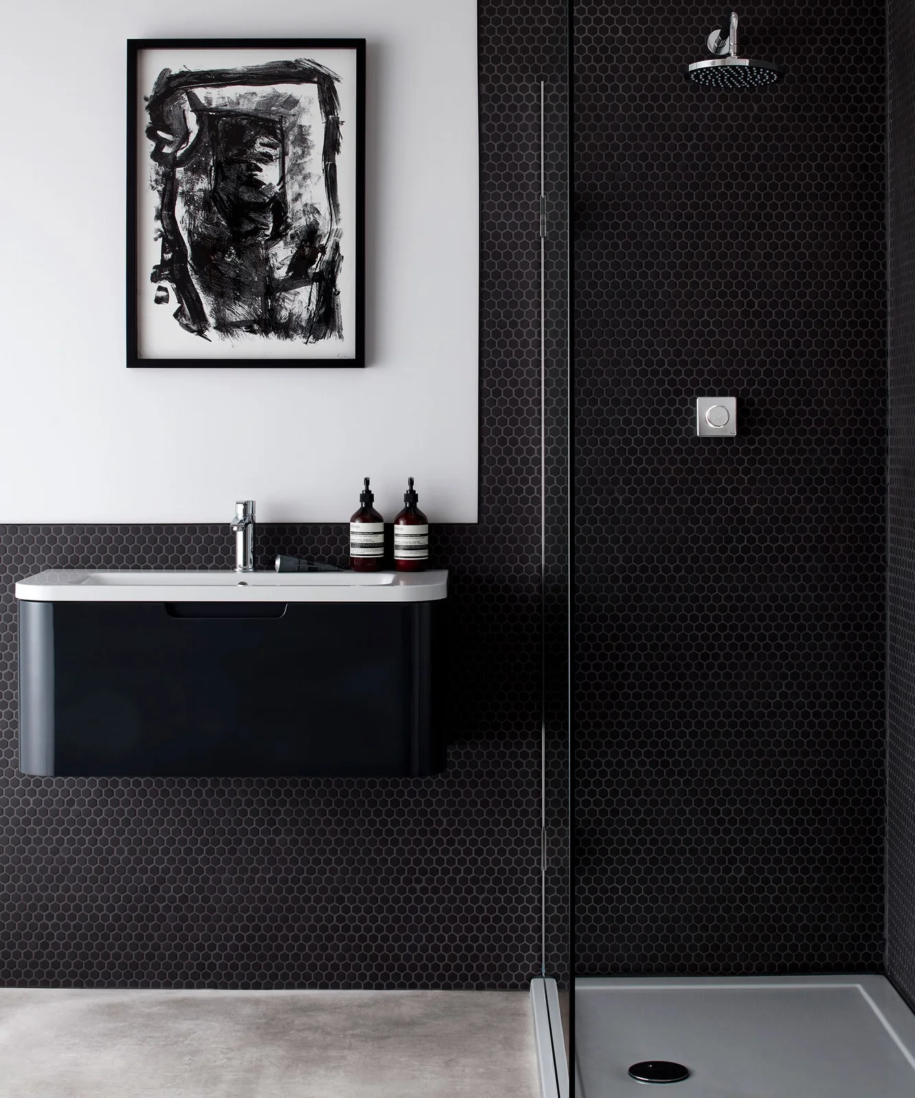 A dark small-tiled shower stands beside a wall-mounted dark vanity and white wall carrying black-framed abstract artwork above a narrow basin