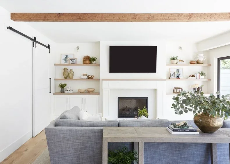 Light living room with gray sofa, white fireplace, television, open wood shelves, lower cupboards and exposed ceiling beam