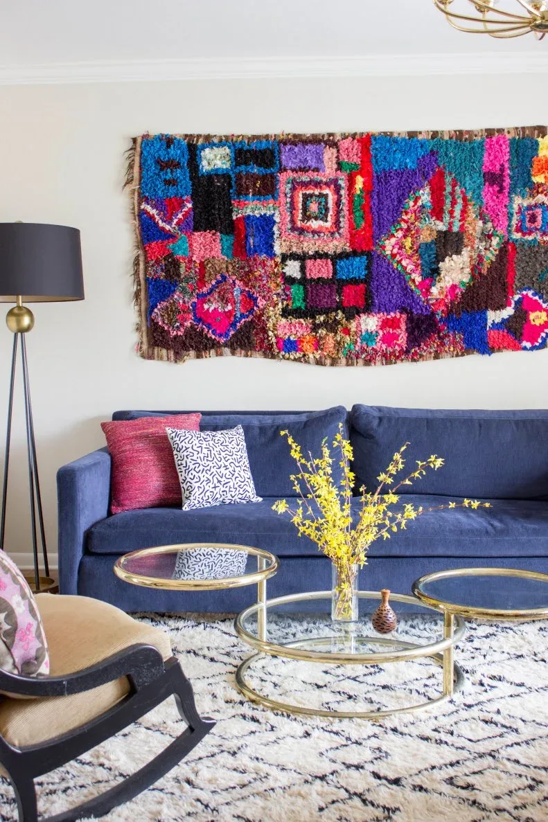 Living room with blue sofa, multicolored geometric textile on pale wall, pink and patterned cushions, round glass tables and cream patterned rug.