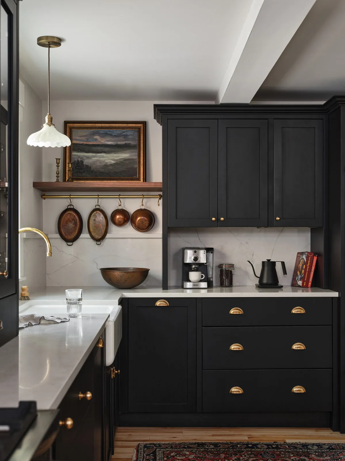 Black drawers with brass cup pulls sit beneath a pale counter, with copper pans hanging below a timber shelf.