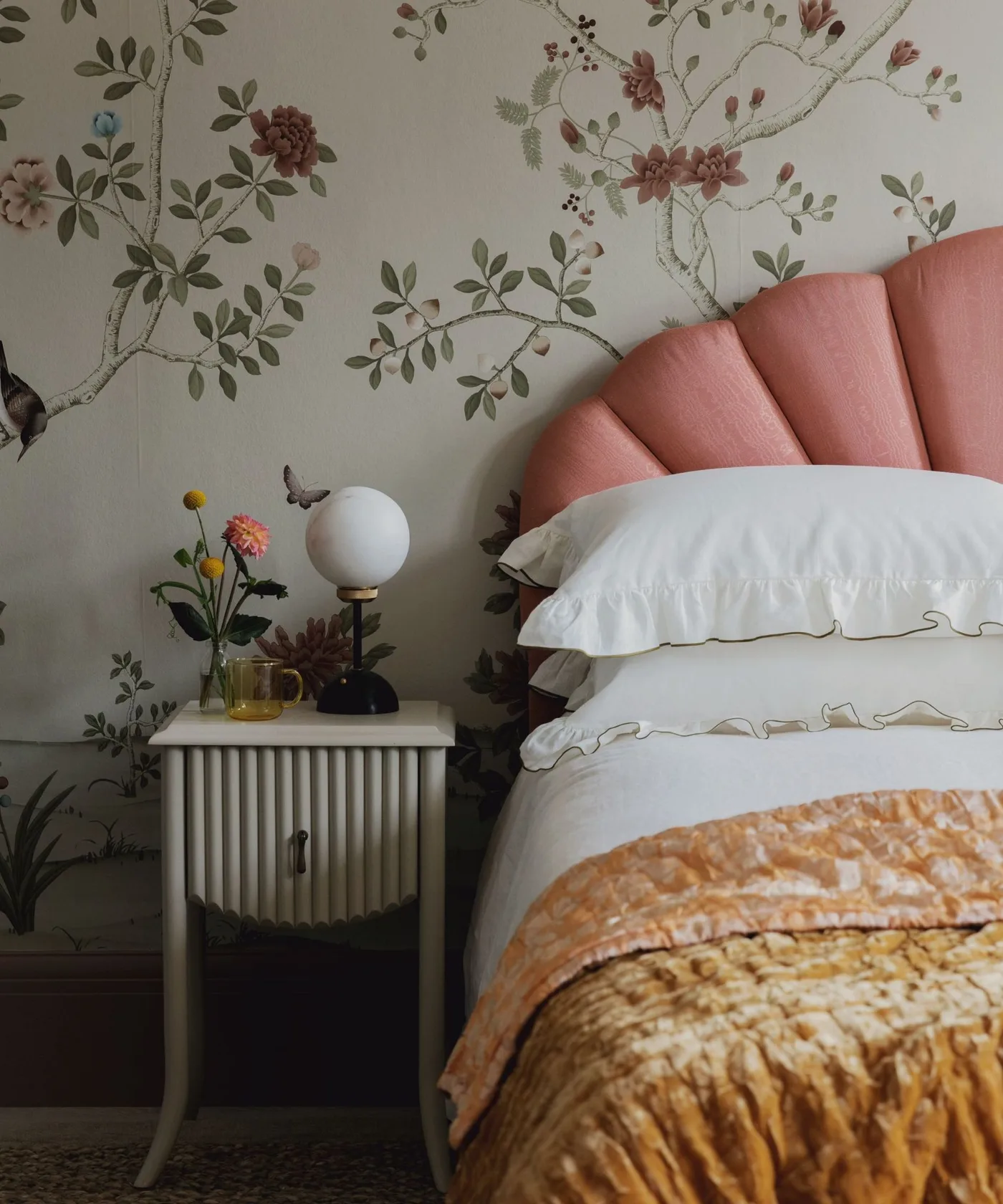 Bedroom detail with a curved pink upholstered headboard, branching floral wallpaper, pale bedding and a small fluted bedside cabinet with a globe lamp.