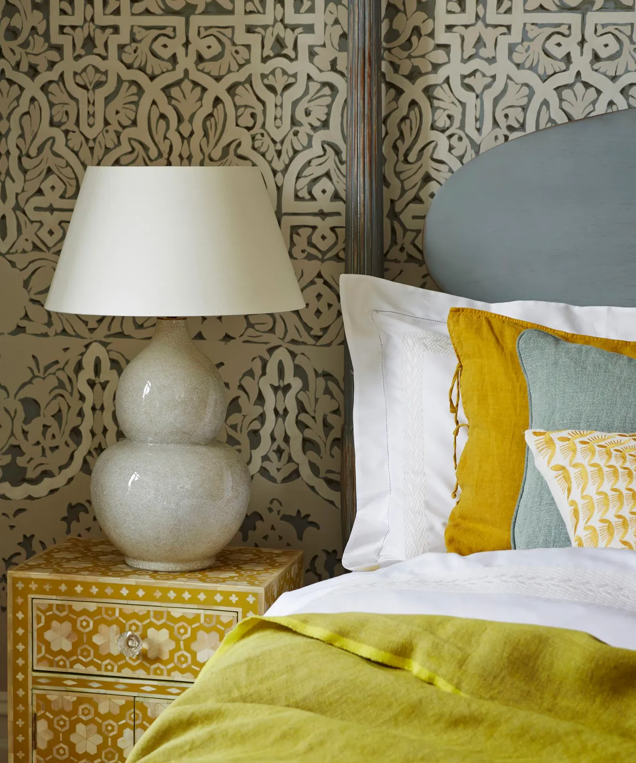Bedroom detail with dark scrolling wall pattern on pale background, curved gray headboard, gray lamp, yellow bedside cabinet and green-and-yellow bedding.