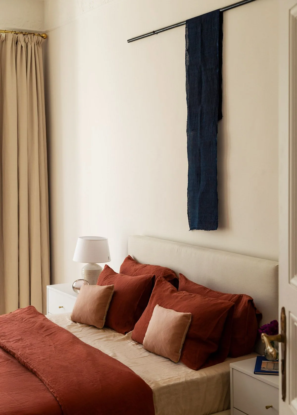 Rust-colored pillows and bed runner in a cream bedroom with pale pink cushions and a narrow dark wall hanging