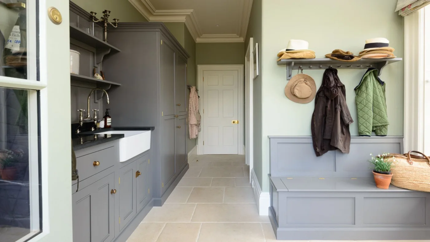 Gray framed cupboards and a white sink stand opposite a pale bench and coat rail beside green walls, large pale floor tiles and a glazed doorway