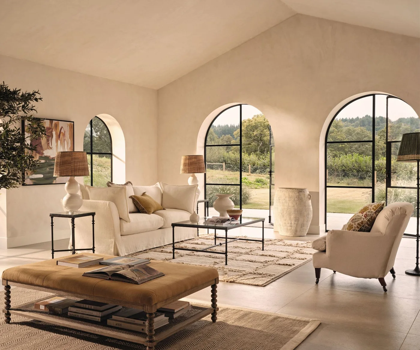 Pale living-room seating and textured rugs sit beside three tall arched openings, slim dark tables, large vessels and a warm-colored upholstered bench