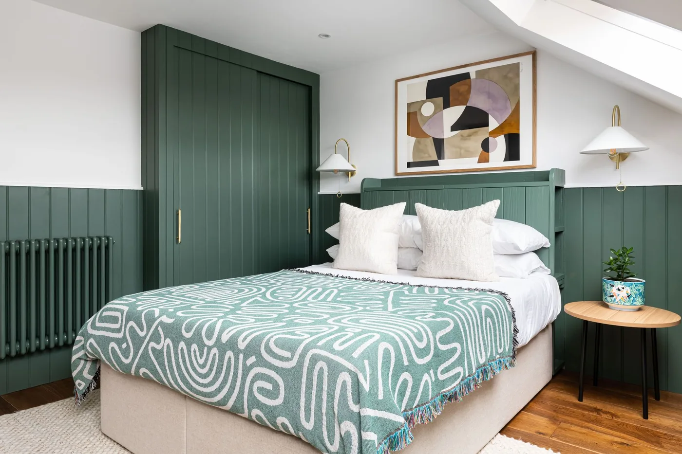 Bedroom with dark green lower paneling and tall wardrobe, white upper walls, pale bed, green patterned blanket, abstract artwork, and white wall lights.