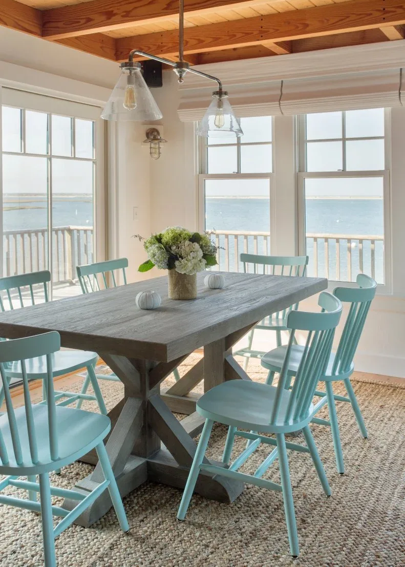 Dining area with gray wooden trestle table, pale blue chairs, woven rug, exposed wooden ceiling and clear hanging lights.