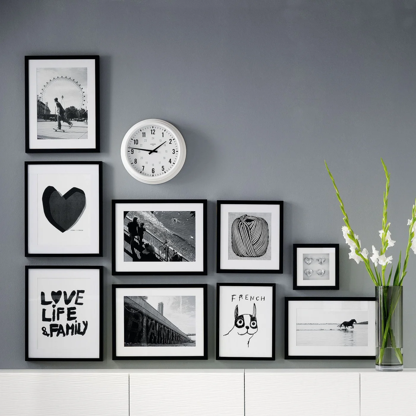 Black picture frames and a white clock form a stepped gallery against a gray wall above a white sideboard