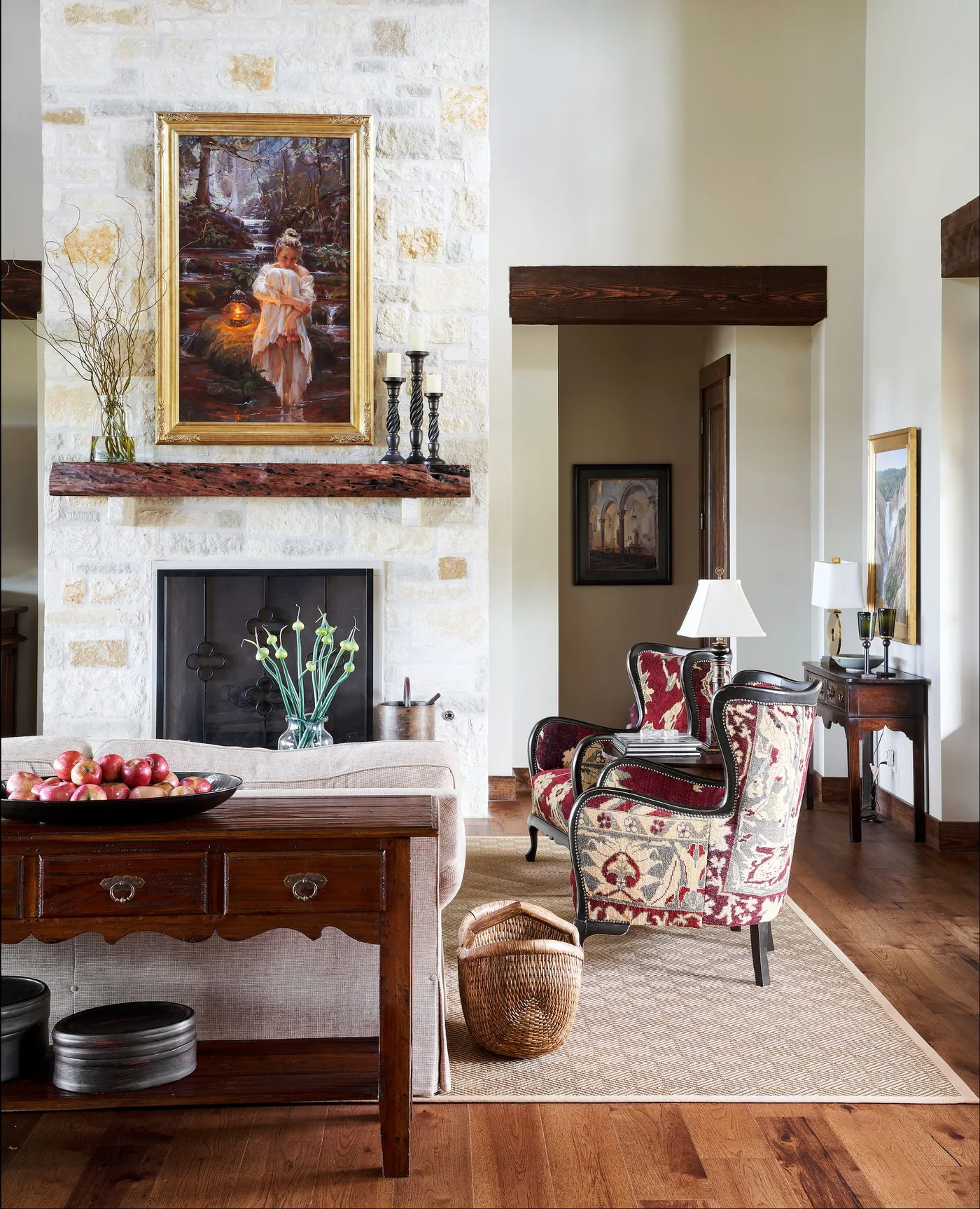 Pale masonry fireplace with reddish wood mantel, framed figure painting, dark candlesticks and patterned chairs beside wood flooring