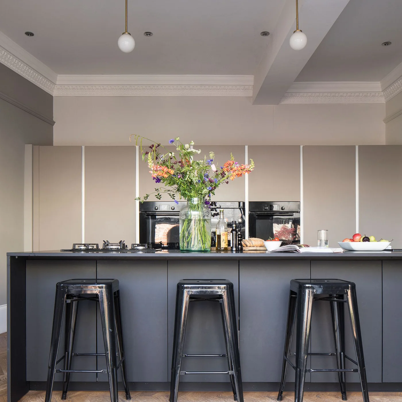 Three dark metal stools sit against a gray flat-front island below two small globe pendants
