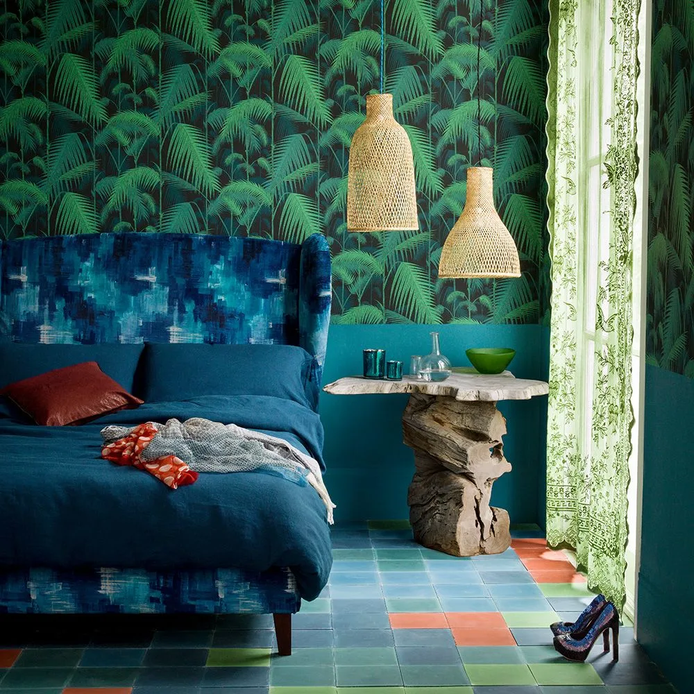 Bedroom with dark green palm wallpaper, patterned teal headboard, deep blue bedding, red cushion, woven pendants, and colorful patterned floor.