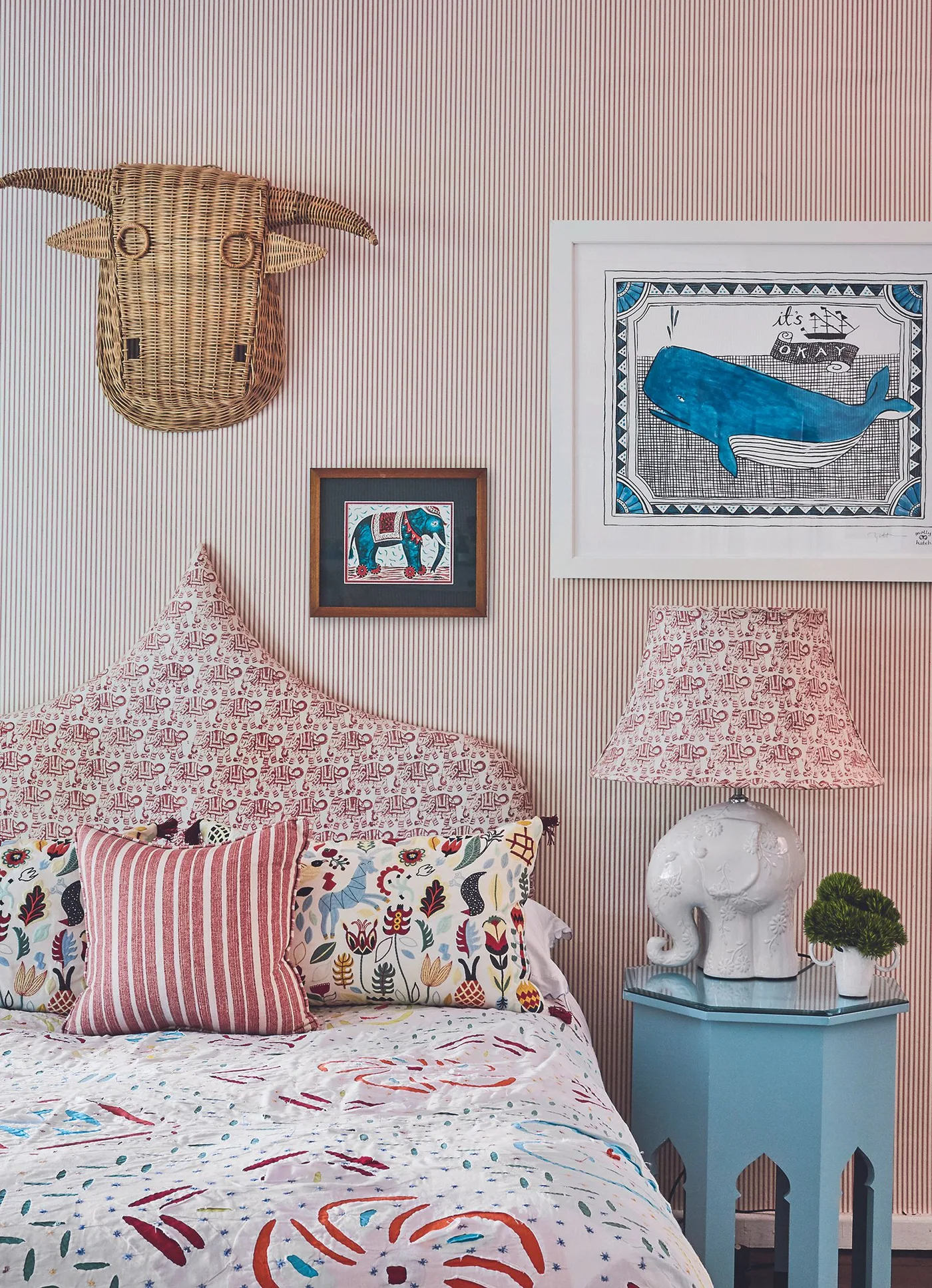 Bedroom detail with pointed patterned headboard, narrow pink wall stripes, printed bedding, blue bedside table, elephant-shaped lamp and woven animal wall decoration.