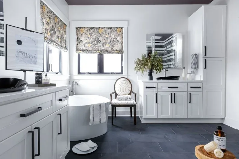 Bathroom with charcoal floor tiles, white cabinets with black handles, black vessel basins, white rounded tub, patterned window blinds, and framed art.