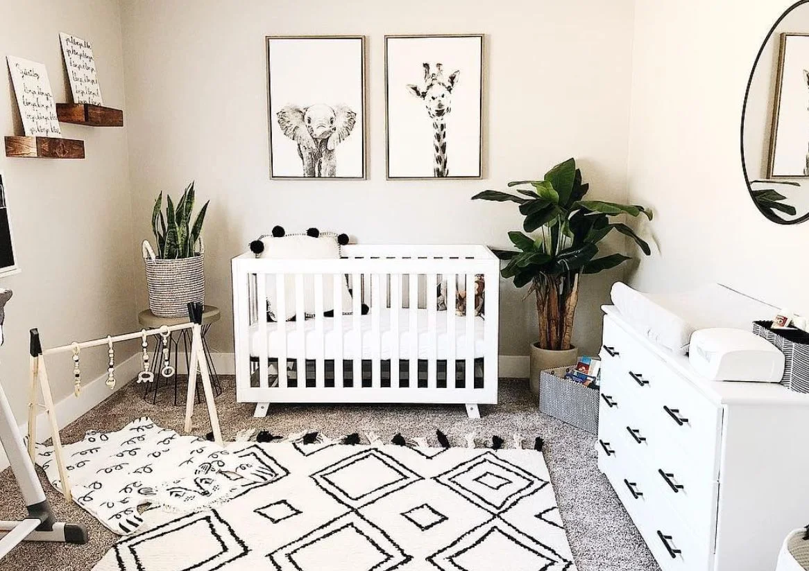A white nursery crib and dresser with black handles sit beside black-framed animal pictures and a large black-and-white geometric rug with plants