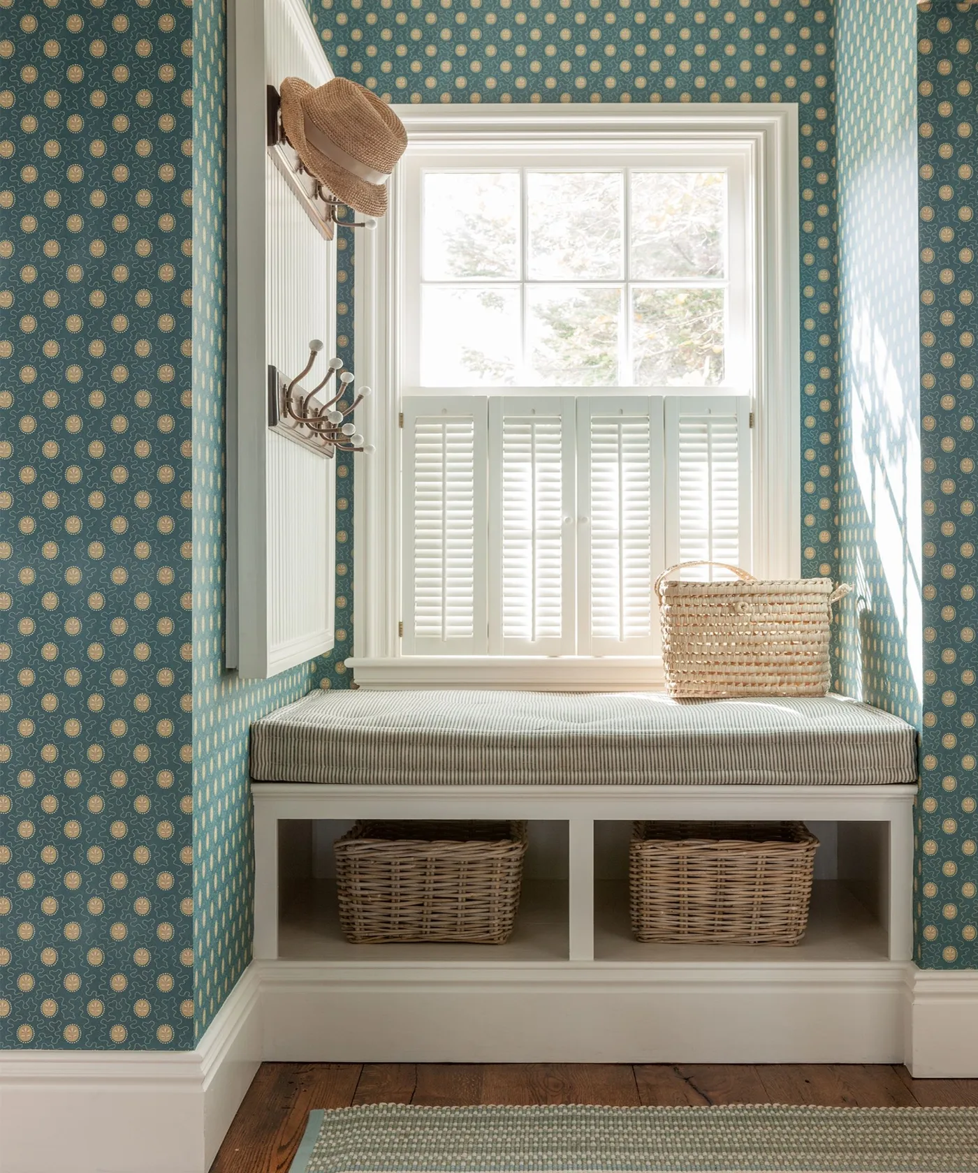 Narrow entry recess with teal dotted wallpaper, cushioned white bench, lower woven baskets, wall hooks and window shutters