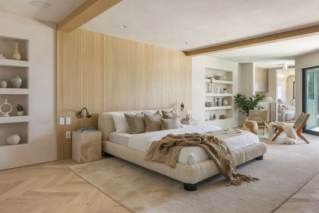 A low pale upholstered bed stands against a light vertically lined wood wall beside open shelves, a pale rug and a light-framed lounge chair