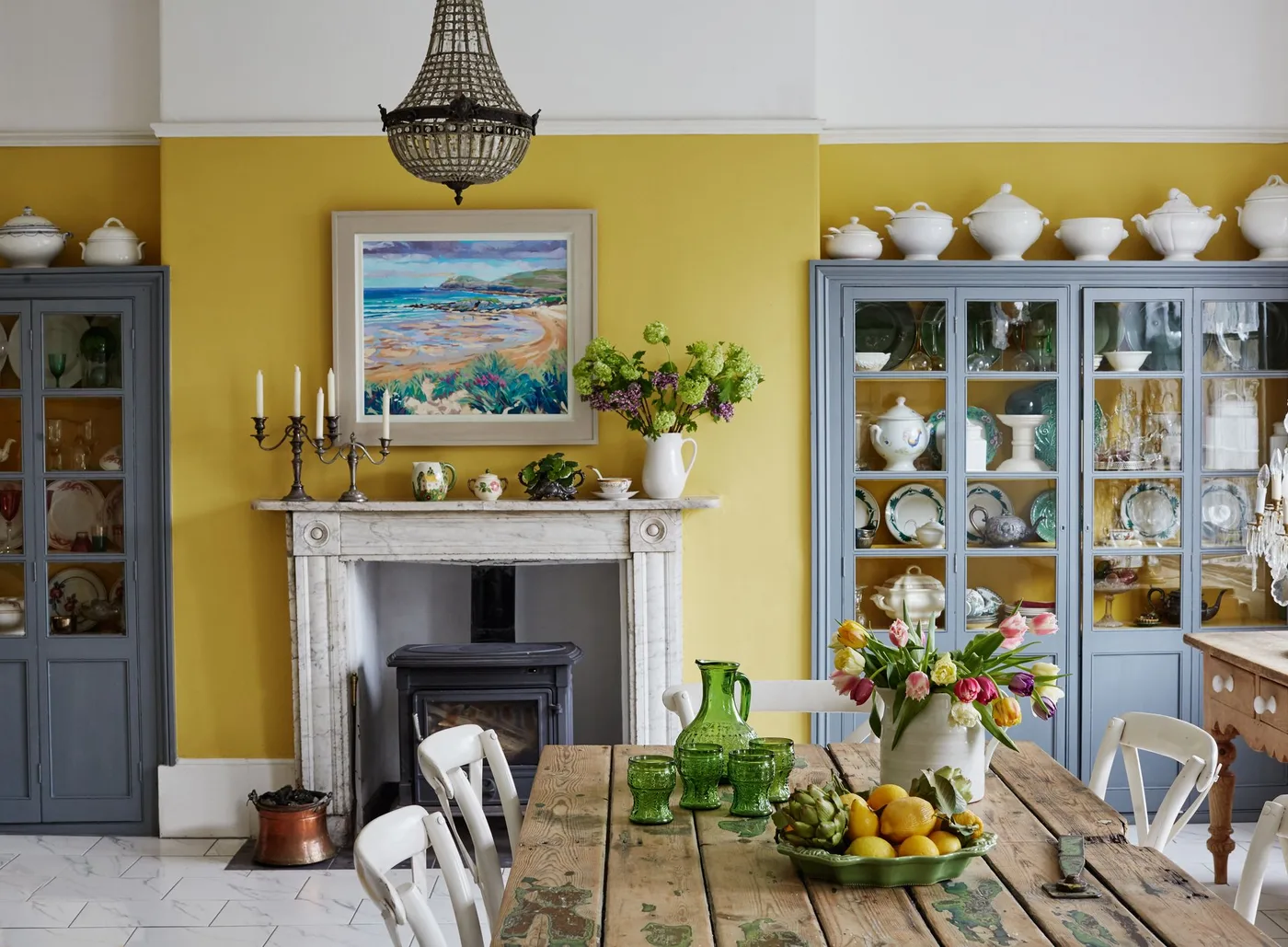 Yellow dining room with pale fireplace, dark stove, copper-toned bucket and glass-front cabinets