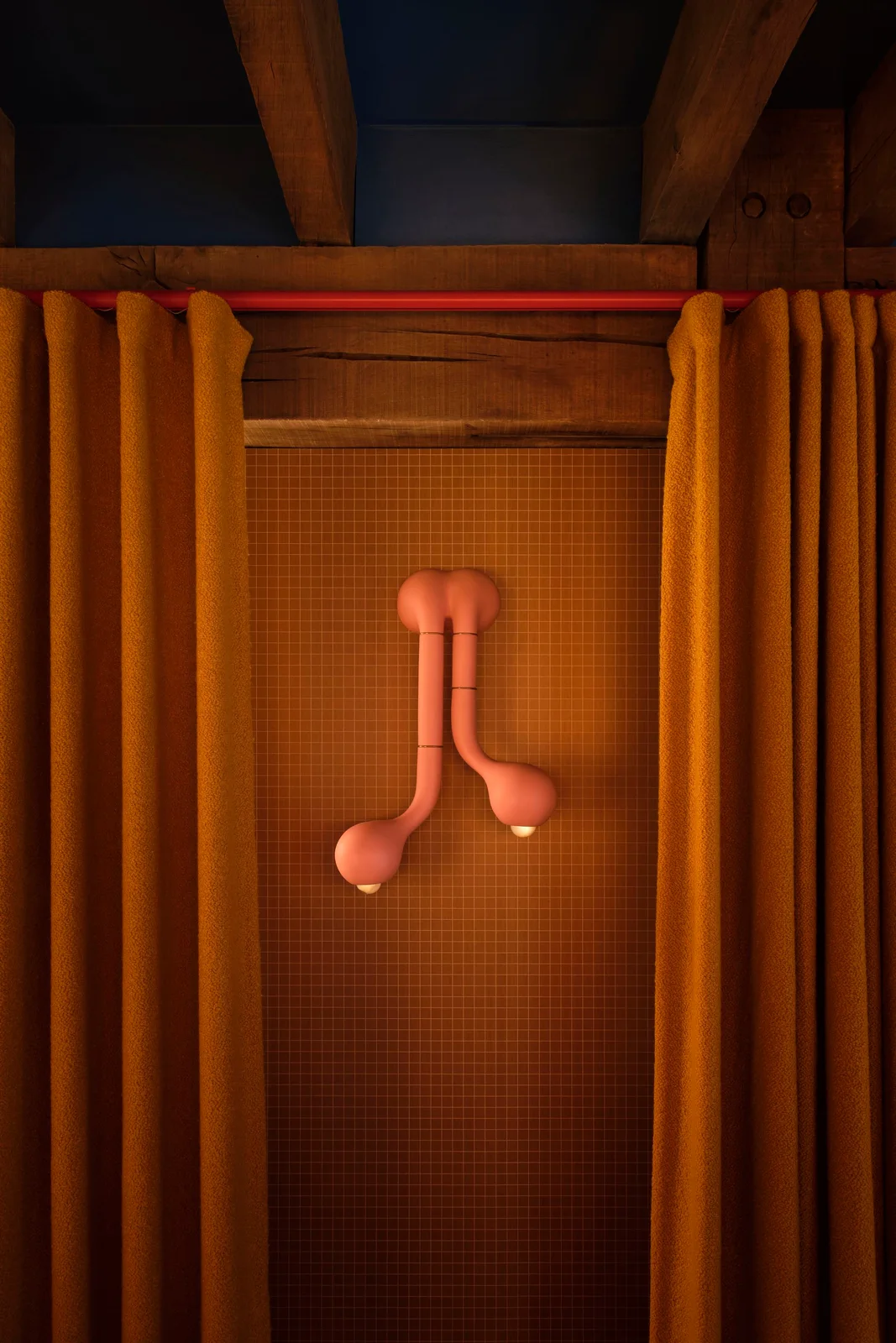 A pink sculptural wall light sits against small warm-colored tiles beneath wooden beams beside yellow curtains on a red rail
