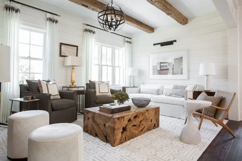 White board-walled living room with exposed beams, pale sofa, darker armchairs, two round stools and block-like wood coffee table
