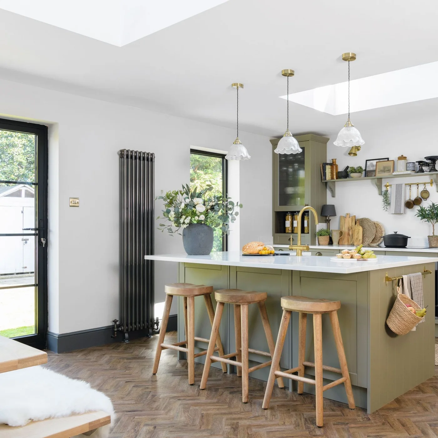An olive green island with a pale counter and wooden stools stands beside light walls and a darker window frame