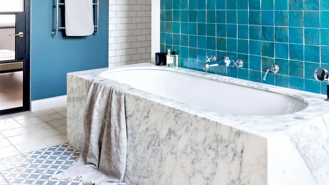A white inset bath has a broad pale veined surround beside blue square wall tiles, a blue painted wall and a small patterned floor section