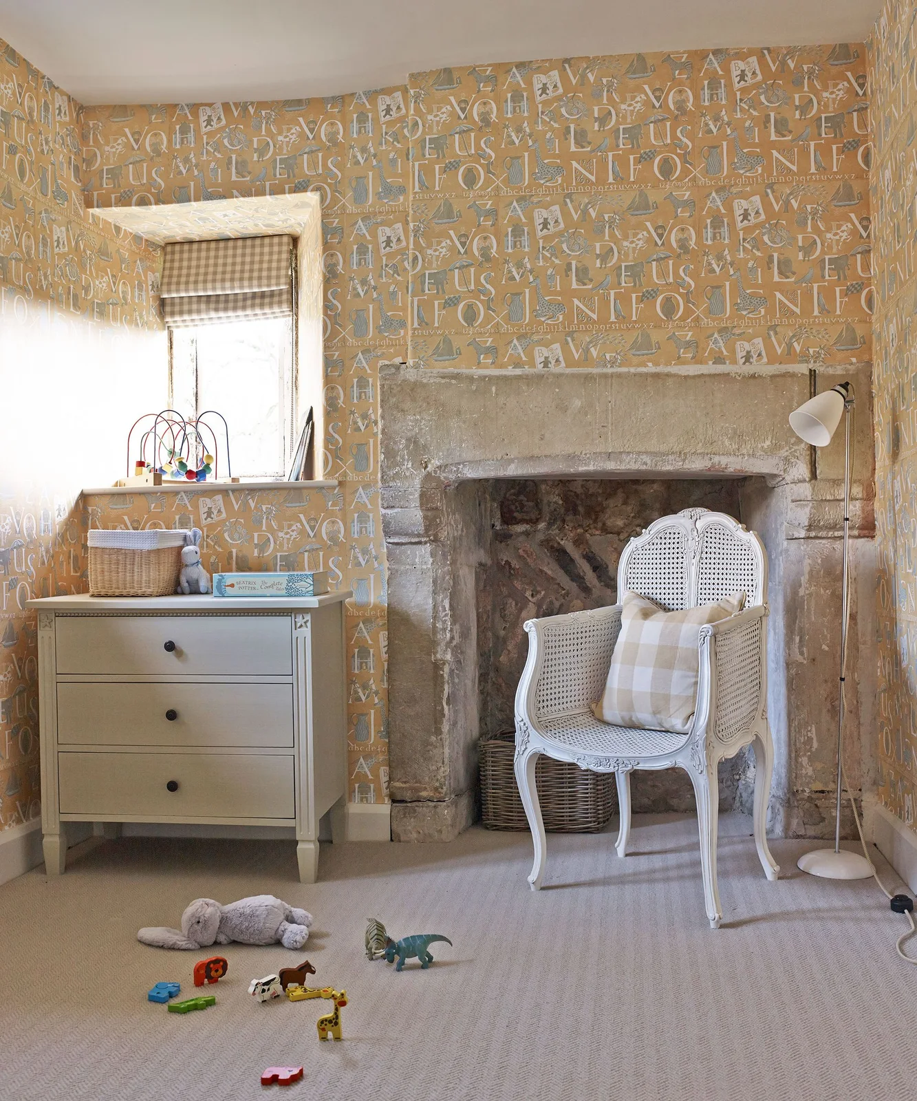Yellow alphabet wallpaper surrounds a checked nursery blind, pale dresser and white woven adult chair beside an old fireplace opening
