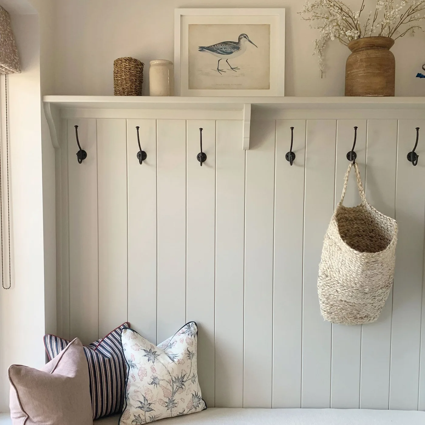 Country entry bench detail with pale vertical wall boards, a row of dark hooks, striped and floral cushions and an upper shelf with a bird picture.