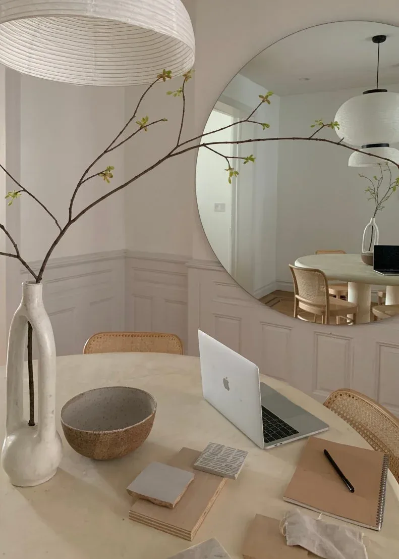 Pale round dining table with a laptop, notebooks, woven-seat chairs and a large round wall mirror