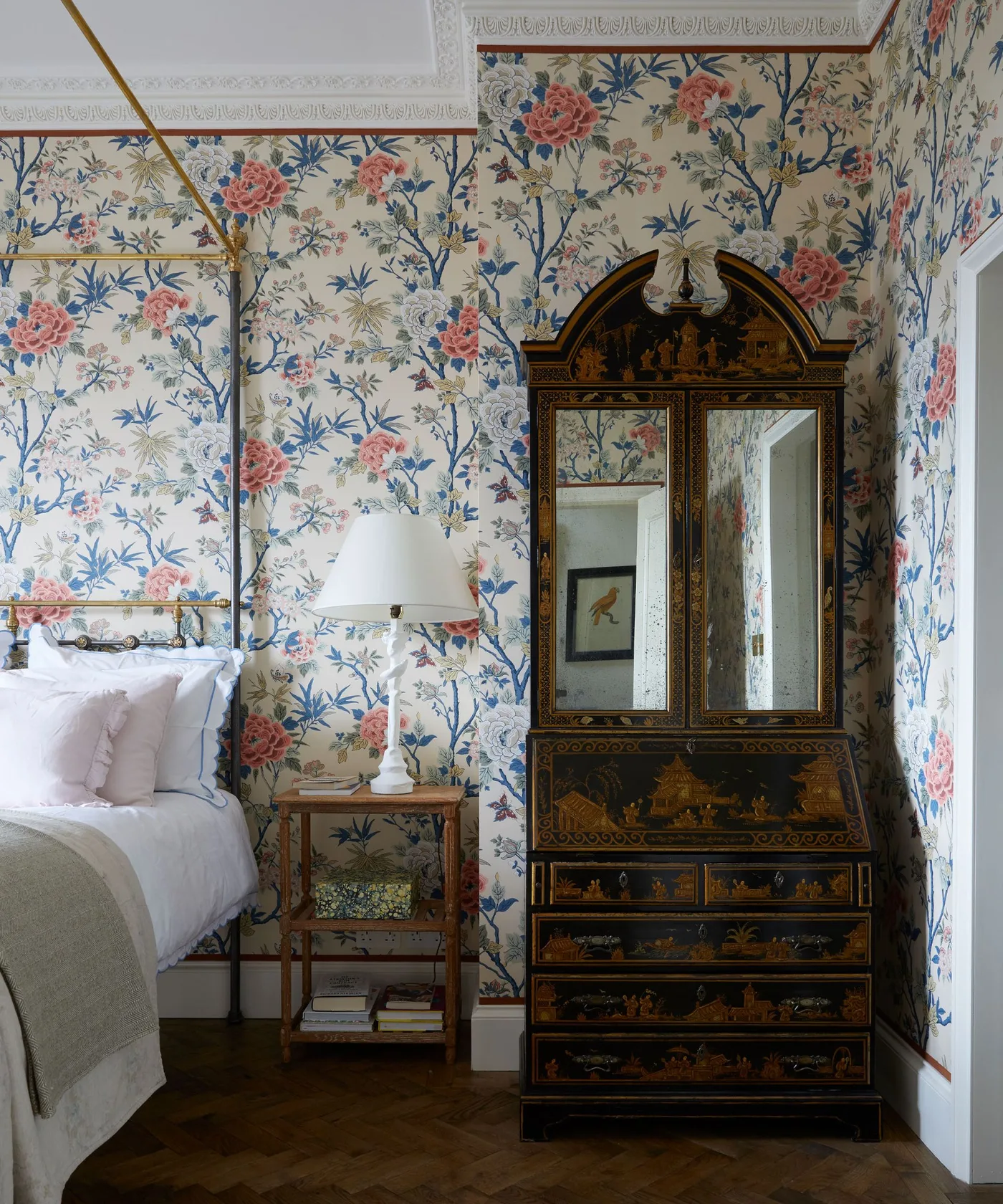 Pink and blue flowers on cream wallpaper beside a slender four-poster bed and a tall decorative cabinet.