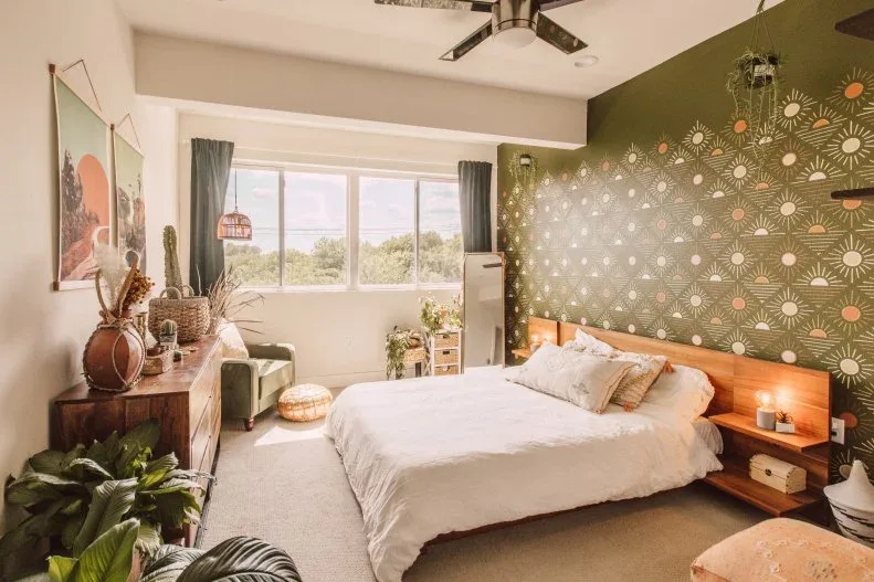 Bedroom with green patterned accent wall, wooden headboard, white bedding, hanging plants, large window and plants beside a wooden dresser.