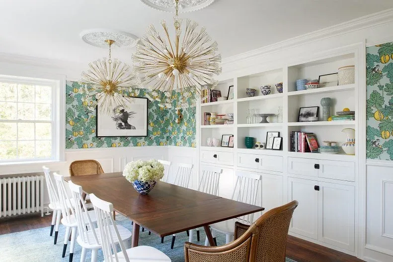 White fitted shelves and lower cabinets beside green floral wallpaper, with a wooden dining table, white chairs, woven end chairs, and starburst lights.