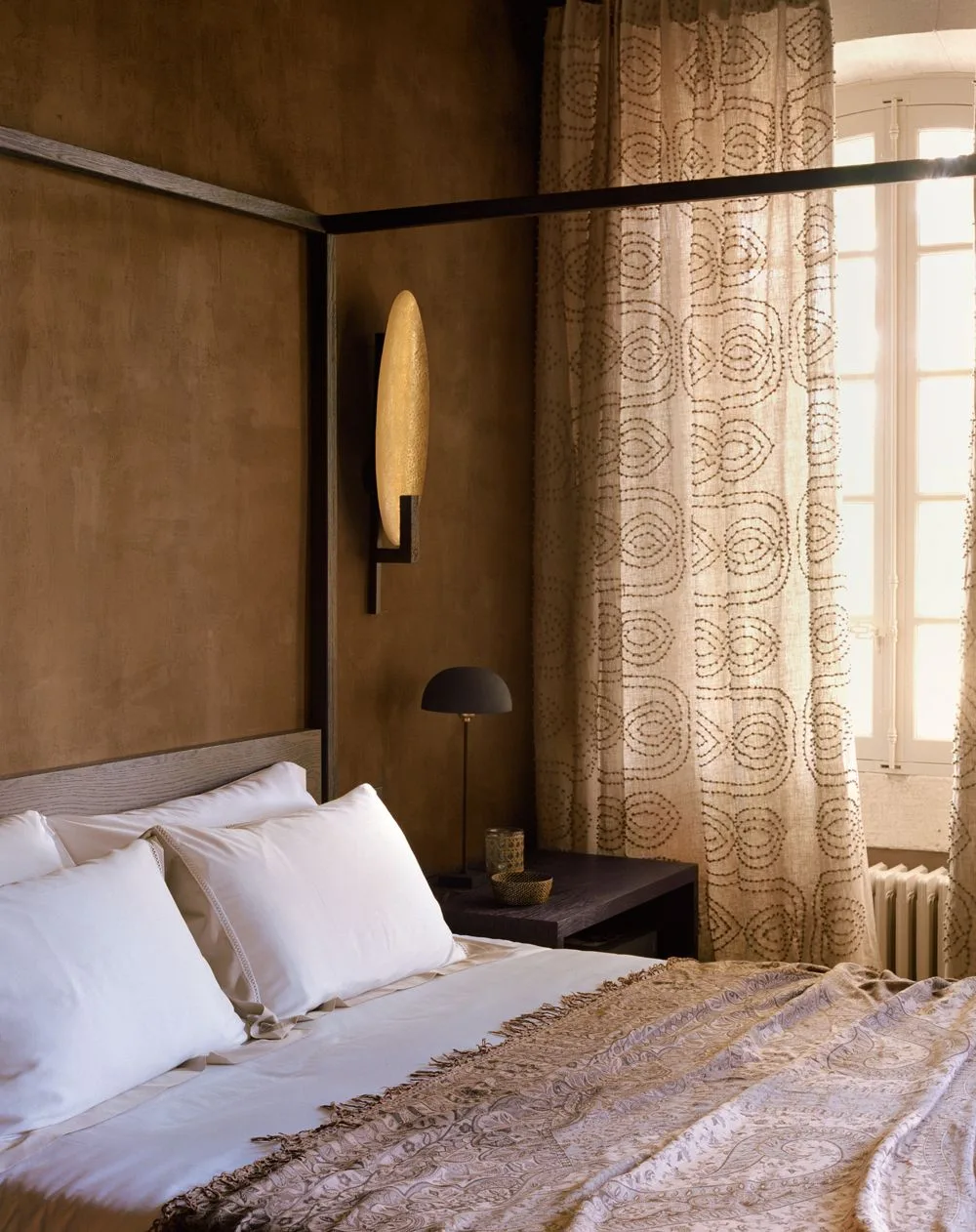 Brown bedroom walls around a dark four-post bed with pale bedding, patterned curtains and a gold-toned wall light