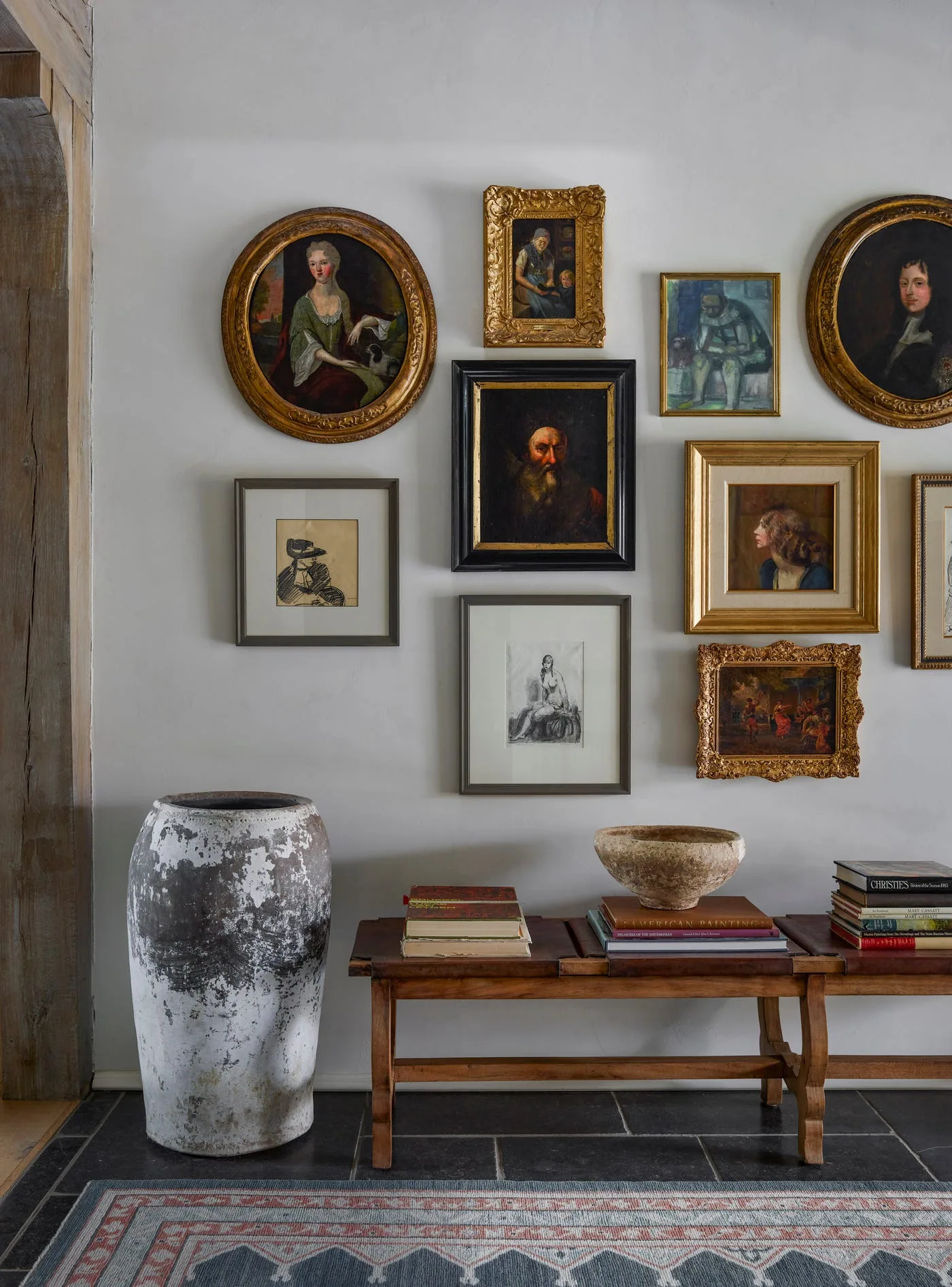 Low wood table with stacked books and bowl beneath mixed portrait frames, beside a large mottled vessel and dark floor tiles