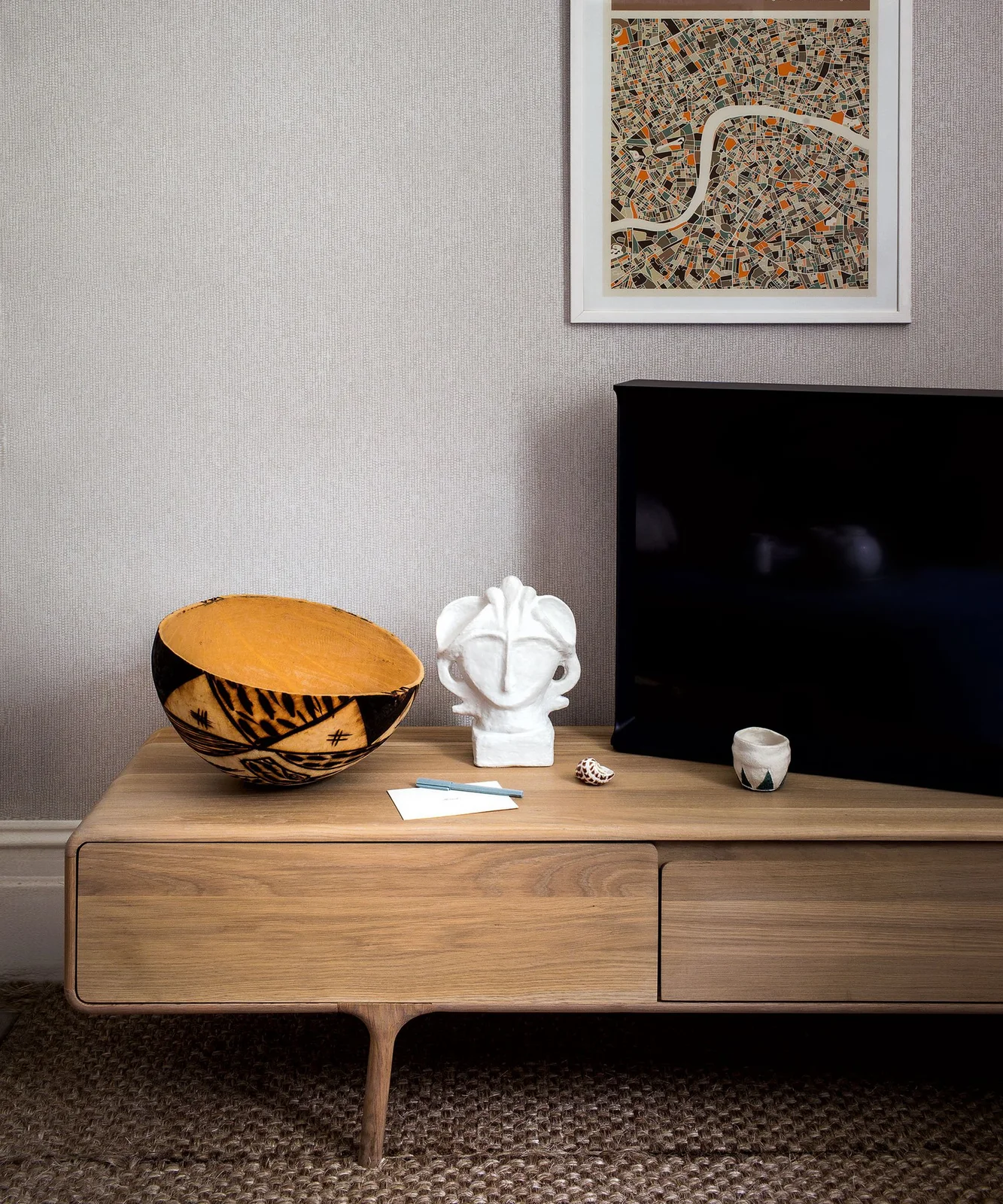 A television stands on a low wooden cabinet beside a yellow patterned bowl and white sculpture beneath framed artwork