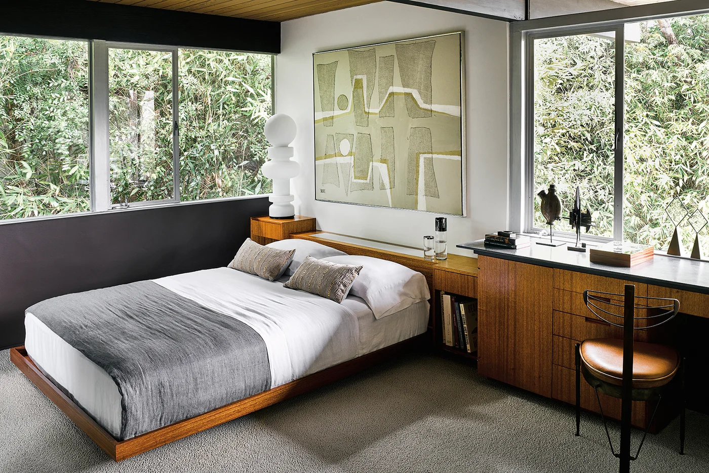 A low wood bedroom bed sits beside a long wood cabinet and desk beneath corner windows with pale bedding, dark lower walls and abstract artwork