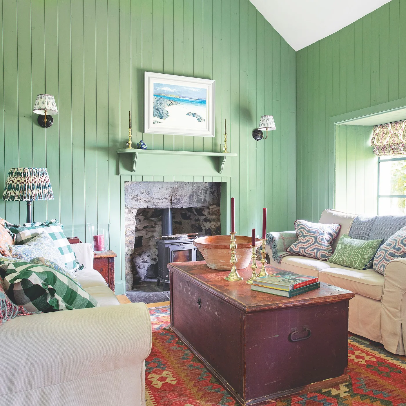Pale sofas face a wooden chest beneath green boarded walls and patterned lamps in a cottage living room