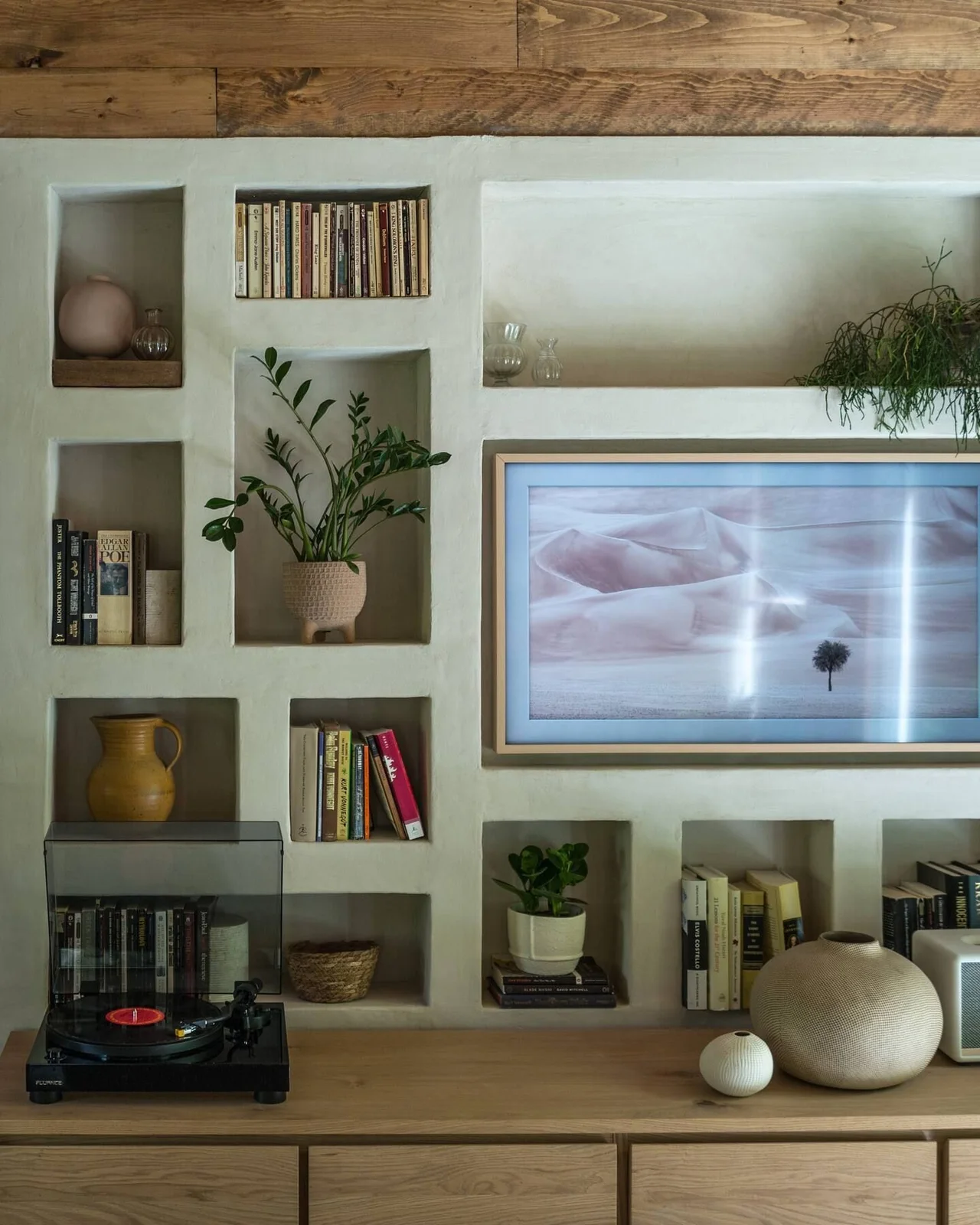 An art-displaying television is set among pale wall recesses holding books, plants and vessels above a wooden cabinet and record player