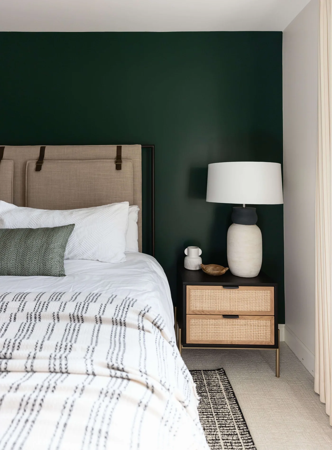 Forest green bedroom wall behind a pale upholstered headboard, white bedding, woven-front bedside drawers, and a large pale lamp.
