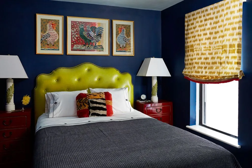 Navy bedroom walls with a bright green tufted headboard, red bedside tables, framed bird artwork, and a yellow patterned blind.